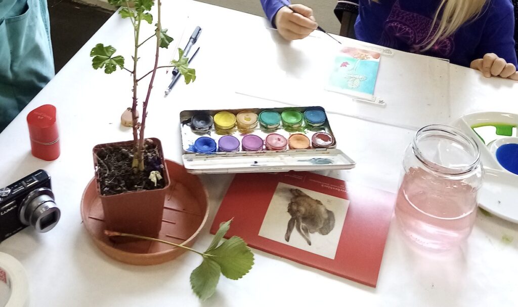 Mesa de taller artístico con acuarelas, una planta, materiales de dibujo y una participante pintando sin mostrar el rostro